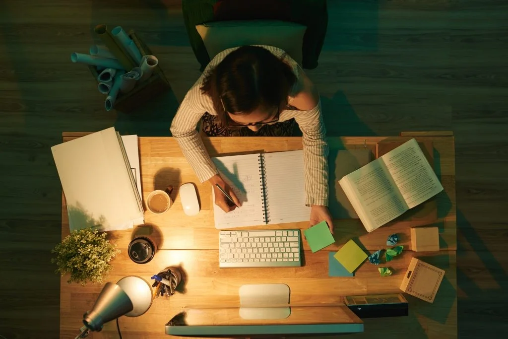 Top-down view of a person working at a well-lit wooden desk in a dark room. They are writing in a spiral notebook with a pen while holding a green sticky note. The desk is cluttered with various items, including a computer monitor, keyboard, mouse, an open book, stacks of paper, two coffee cups, a small potted plant, and colorful sticky notes. A desk lamp on the left provides warm light, casting shadows across the workspace. In the corner, a bin holds several rolled-up blueprints or posters.